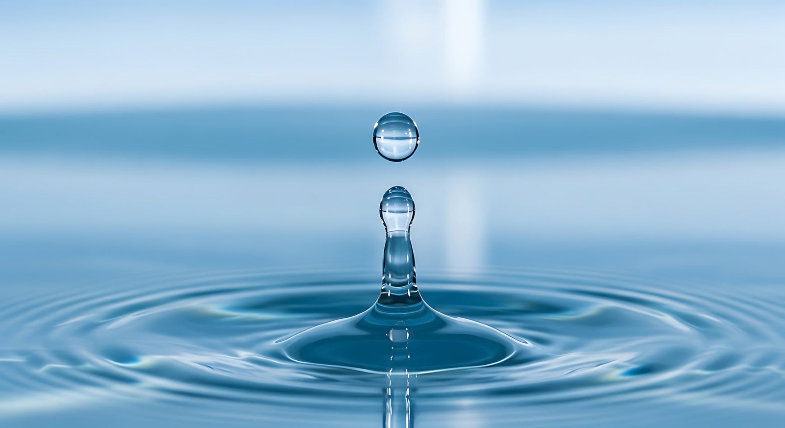 A close-up photograph of a single clear water droplet hanging above a water fountain, with ripples spreading across blue water under soft light.